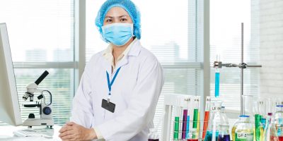 Portrait shot of confident Asian microbiologist wearing white coat and medical mask sitting at desk and posing for photography, interior of modern laboratory on background